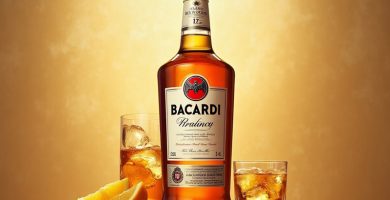Botella de ron Bacardí con dos vasos con hielo y rodajas de naranja sobre fondo dorado